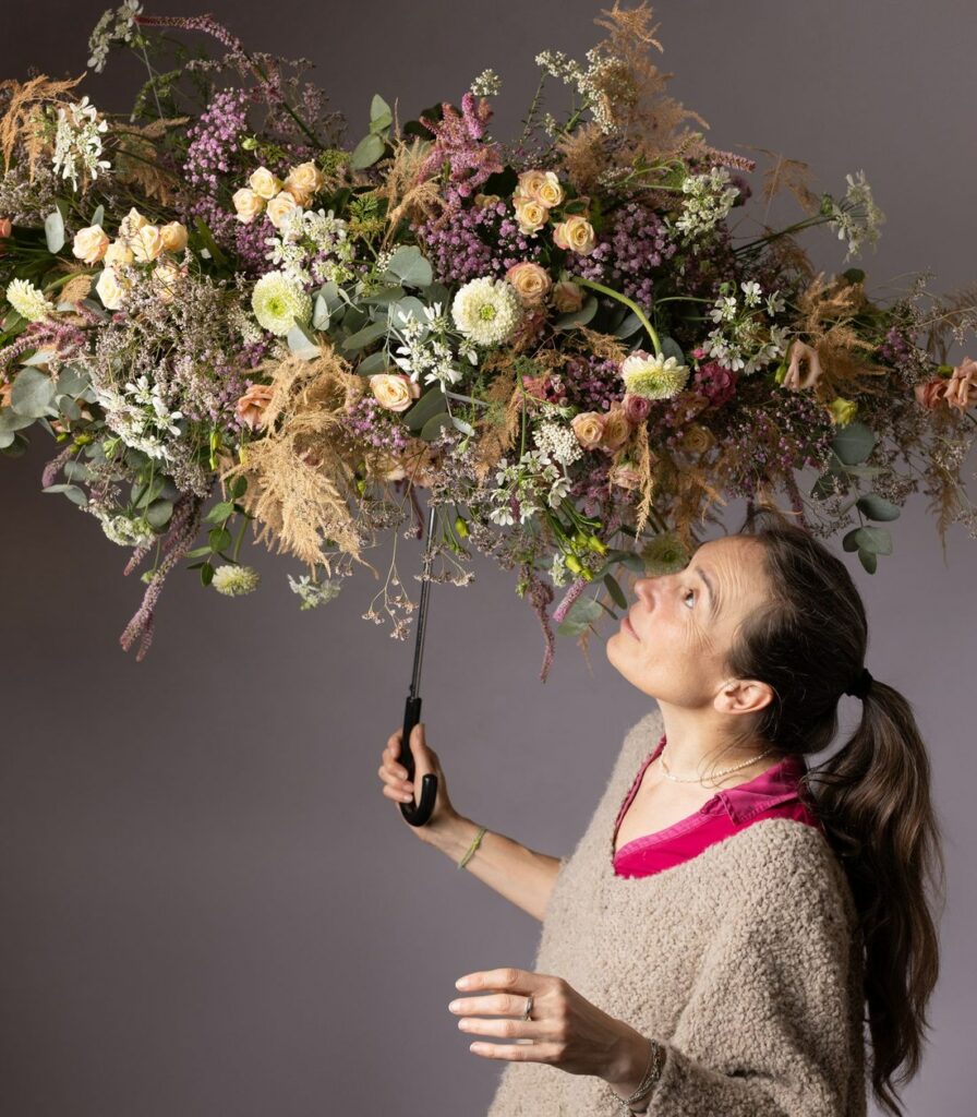 Une fleuriste, tête levée, tient sous elle un parapluie ouvert entièrement fleuri de roses claires, de baies et de graminées séchées.