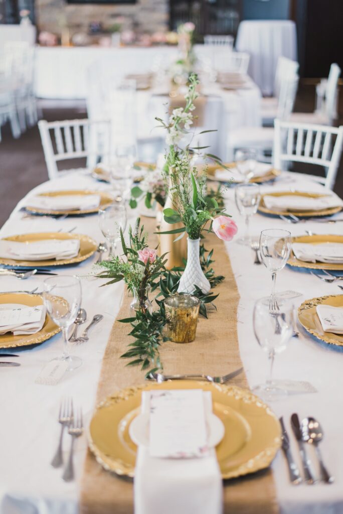 Longue tablée de réception, chemin de table en toile de jute, assiettes de présentation dorées et soliflores garnis d'eucalyptus et de roses pâles.