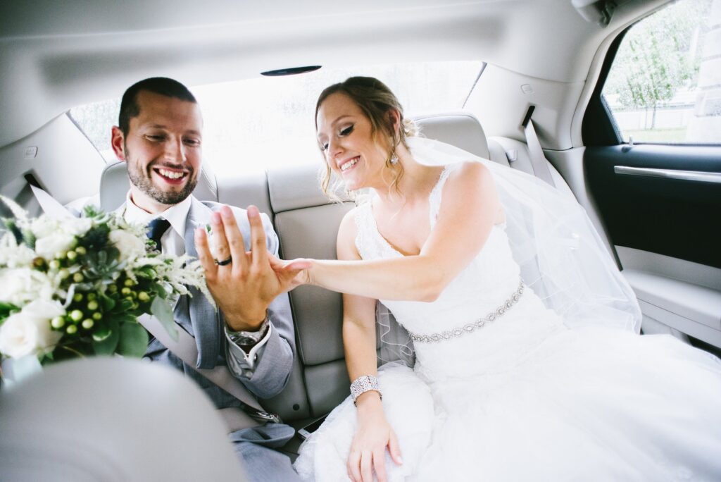 Les mariés assis à l'arrière d'une voiture, main dans la main et riant, le bouquet de roses blanches posé à côté d'eux.
